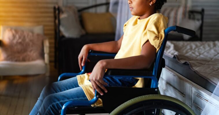 Side portrait of disabled little girl sitting in a wheelchair at home.