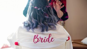 A bride gets her make-up done. She is not facing the camera, the back of her robe reads bride in red letters.