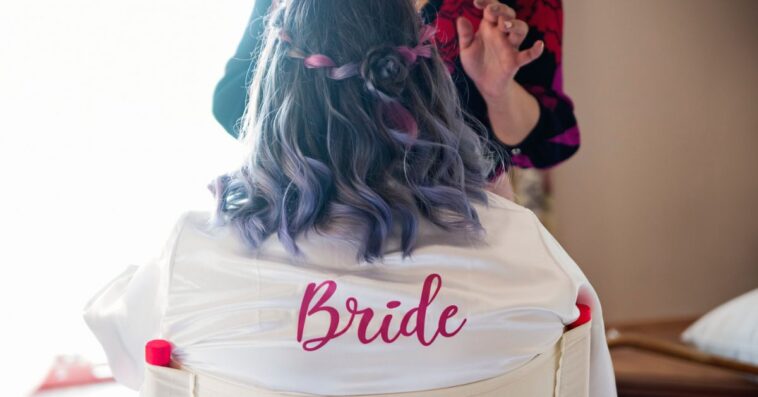 A bride gets her make-up done. She is not facing the camera, the back of her robe reads bride in red letters.