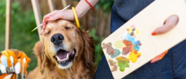 A young female artist works on her art canvas painting outdoors in her garden with golden retriever keeping her company.