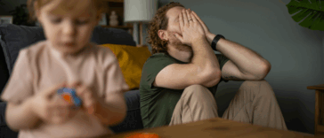 A man leaning against the sofa with his head in his hands and a child playing in front of him.