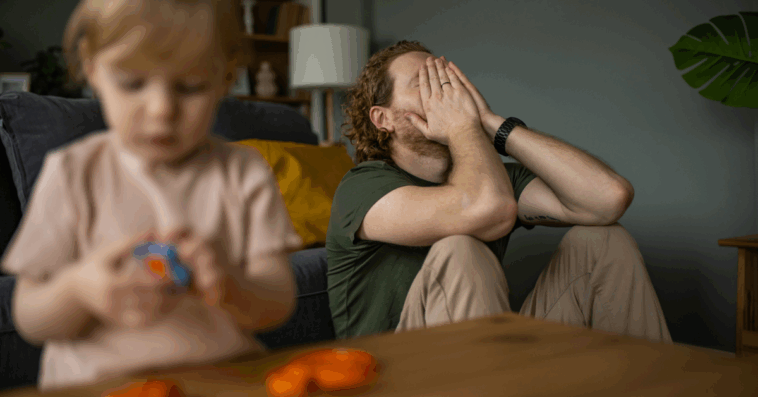 A man leaning against the sofa with his head in his hands and a child playing in front of him.