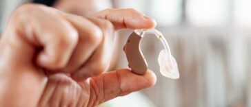 Close-up of senior hand holding a behind-the-ear hearing aid indoors.