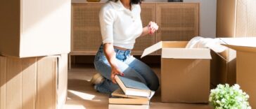 Beautiful woman unpacking her favorite books in her new apartment. Kneeling on the floor taking books out of cardboard boxes to place on the living room shelf.