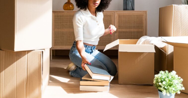 Beautiful woman unpacking her favorite books in her new apartment. Kneeling on the floor taking books out of cardboard boxes to place on the living room shelf.