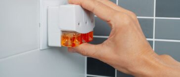 A man's hand plugs a plug-in air freshener into a socket on a tiled bathroom wall.