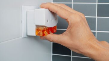 A man's hand plugs a plug-in air freshener into a socket on a tiled bathroom wall.