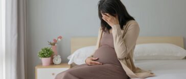 Side view of a pregnant woman touching her belly and sitting on a bed - she is feeling depressed and sad, crying in a bedroom.