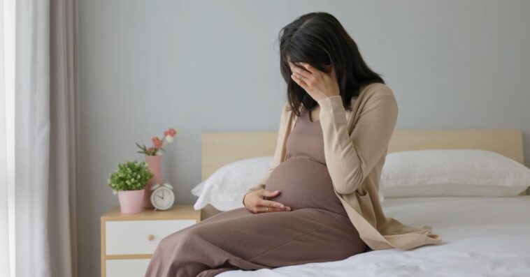 Side view of a pregnant woman touching her belly and sitting on a bed - she is feeling depressed and sad, crying in a bedroom.