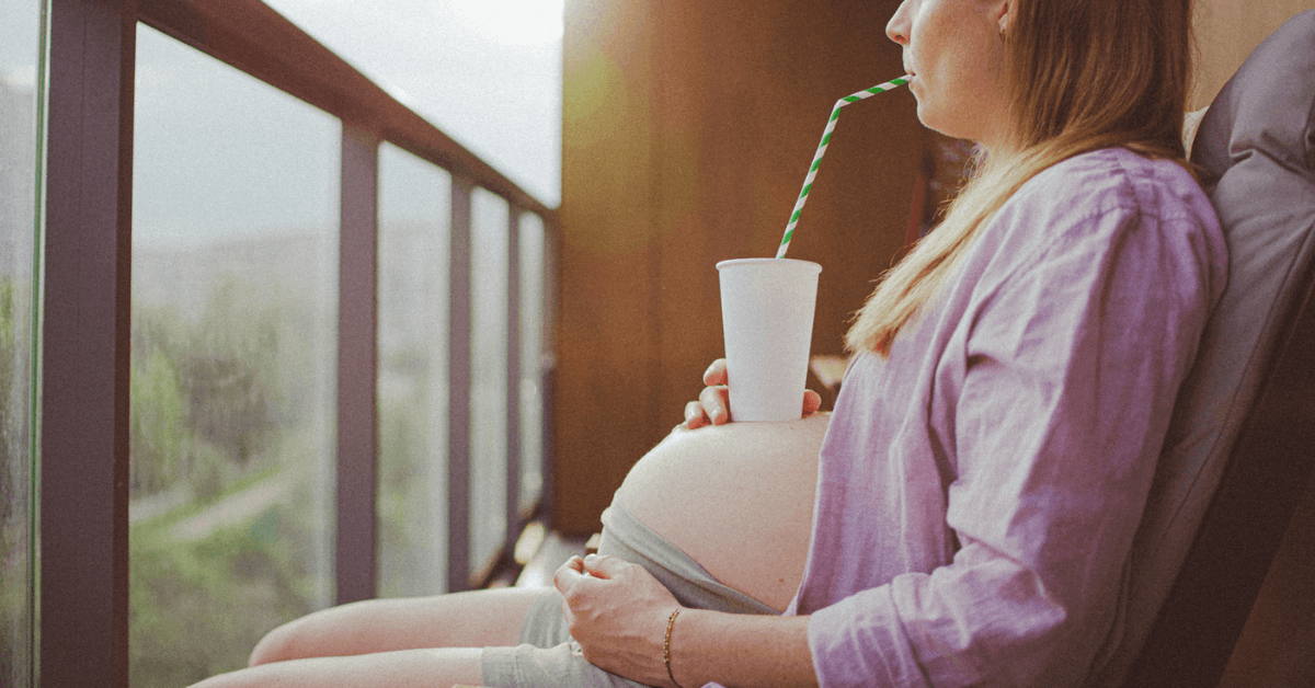 A pregnant woman drinking from a cup resting on her baby bump.