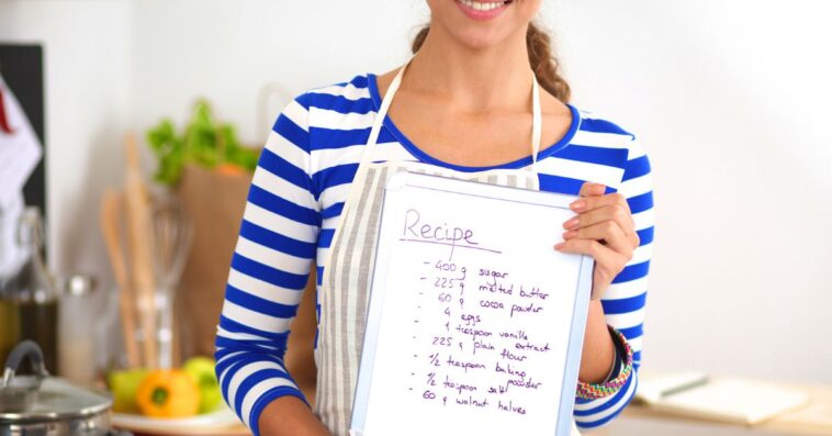 Woman in the kitchen at home, standing near stove while holding a recipe.