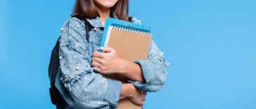 Happy cute teenage girl wearing oversized denim jacket, black eyeglasses and backpack holding books. Smiling teenager standing against blue background.