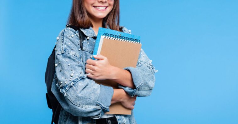 Happy cute teenage girl wearing oversized denim jacket, black eyeglasses and backpack holding books. Smiling teenager standing against blue background.