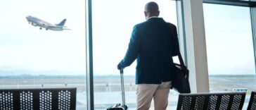 Airport, suitcase and businessman waiting to board flight by the terminal for corporate work trip. Travel, luggage and professional male traveler watching flying plane by a window in terminus lounge.