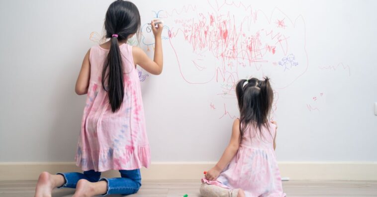 Back shot of two young girls drawing with markers on a white wall.
