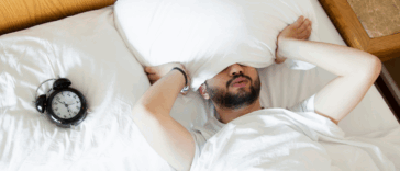 person laying on bed with head under pillow with alarm clock next to them