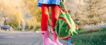 feet and legs of child in pink rainboots holding a green backpack