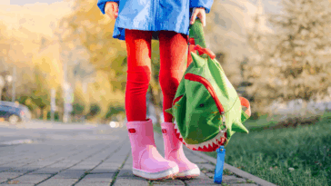 feet and legs of child in pink rainboots holding a green backpack