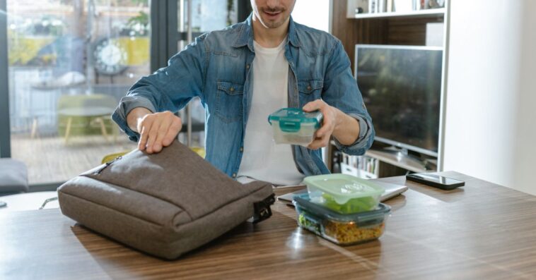 A man organizes lunch boxes while preparing for his day. The background shows a modern living space. This action emphasizes preparation, organization, and a healthy lifestyle.