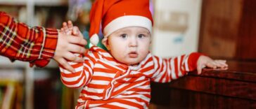 Baby in Christmas pajamas and Santa hat at home with sibling sister by Christmas tree.