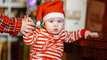 Baby in Christmas pajamas and Santa hat at home with sibling sister by Christmas tree.