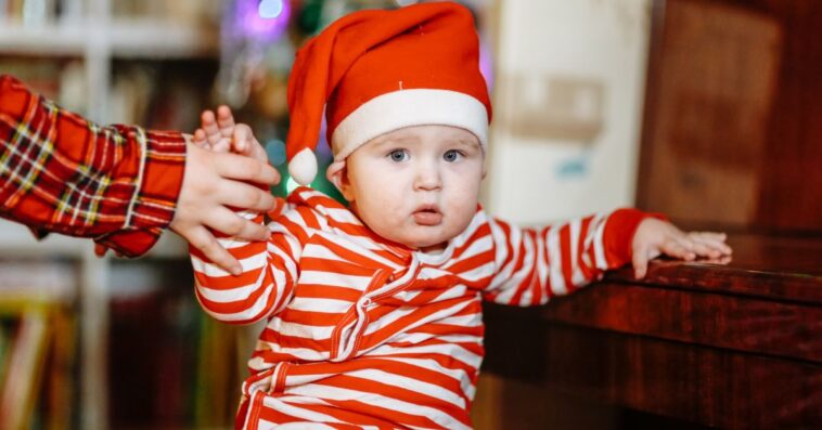 Baby in Christmas pajamas and Santa hat at home with sibling sister by Christmas tree.
