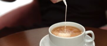 In the cozy teahouse. Male hand pouring milk into coffee in a white ceramic cup on saucer. Closeup of natural hot beverage. Front view, high angle.