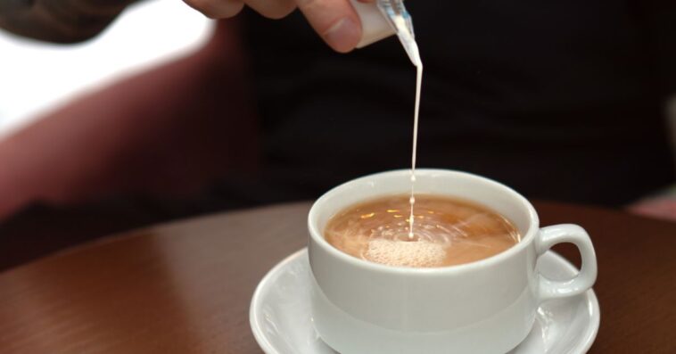 In the cozy teahouse. Male hand pouring milk into coffee in a white ceramic cup on saucer. Closeup of natural hot beverage. Front view, high angle.