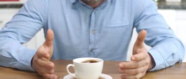 Unidentifiable man brewing coffee at home spills his coffee, looking at camera with sad and confused expression.