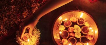 A close-up view of a woman’s hand placing a lit clay lamp (diya) surrounded by marigold flowers, creating a warm and serene ambiance during Diwali, the Hindu festival of lights. This traditional ritual symbolizes the victory of light over darkness.