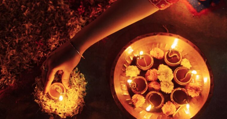 A close-up view of a woman’s hand placing a lit clay lamp (diya) surrounded by marigold flowers, creating a warm and serene ambiance during Diwali, the Hindu festival of lights. This traditional ritual symbolizes the victory of light over darkness.