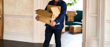 A smiling, mature man walking into empty room of new home, carrying a cardboard box and looking around happily. Excitement, new home, moving, house.