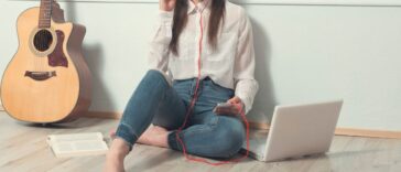Young woman sitting on wooden floor listening to music through headphones. Next to her are a laptop, a smartphone and a guitar.