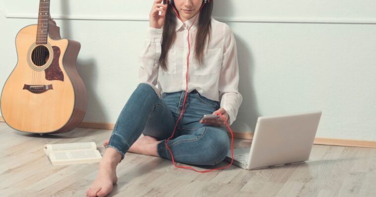 Young woman sitting on wooden floor listening to music through headphones. Next to her are a laptop, a smartphone and a guitar.