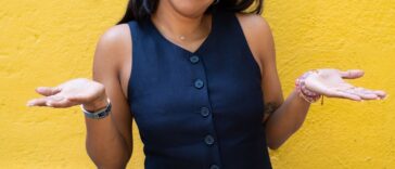 A woman standing against a yellow wall shrugs her hands and head to the side.