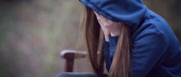 A teenage girl in hooded top, with head in hands in despair. She sits on a park bench.