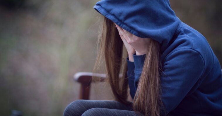 A teenage girl in hooded top, with head in hands in despair. She sits on a park bench.