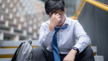 A young, anxious businessman with his head in his hands, sitting outside his building.