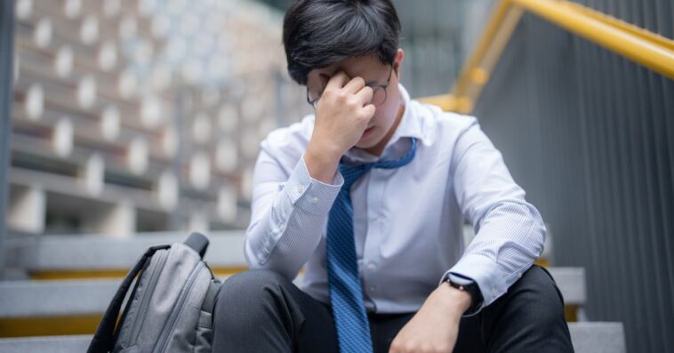 A young, anxious businessman with his head in his hands, sitting outside his building.