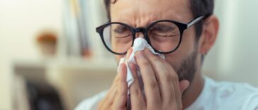 Young man with glasses sneezing, wiping his nose with a piece of tissue paper.