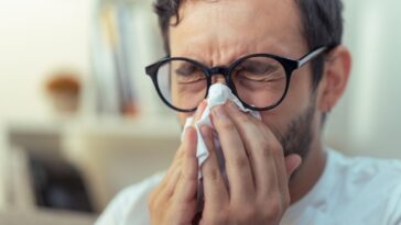 Young man with glasses sneezing, wiping his nose with a piece of tissue paper.