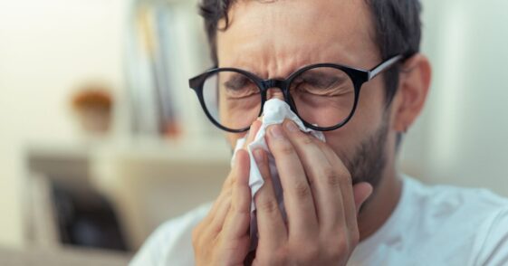 Young man with glasses sneezing, wiping his nose with a piece of tissue paper.