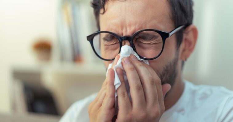 Young man with glasses sneezing, wiping his nose with a piece of tissue paper.