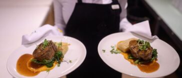 Waiter holding hot plated grilled steak and vegetables potatoes asparagus butter nut garnish sauce.