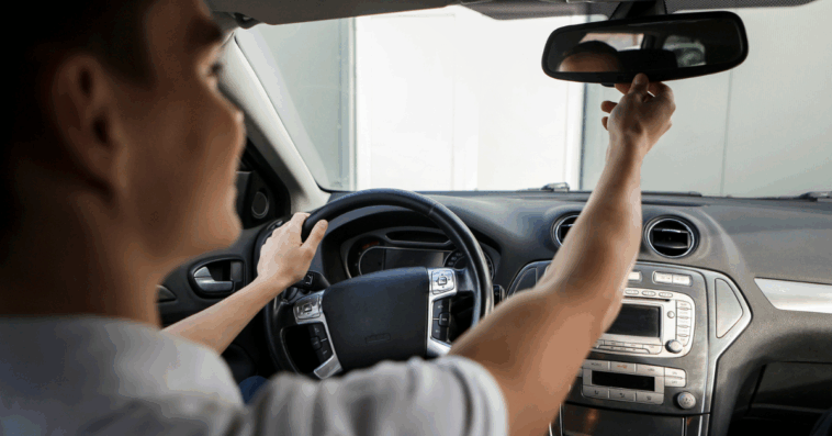 A teenager driving and adjusting their rearview mirror.