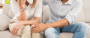 A woman sitting on the sofa crying with a man sitting next to her comforting her.