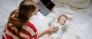 A woman lying on the bed in front of a laptop with a baby by her side.