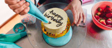 A woman writing happy birthday on a cake with a pastry bag.