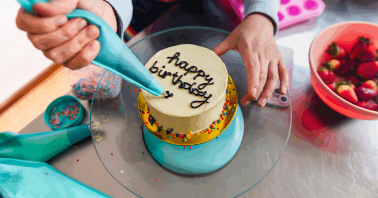 A woman writing happy birthday on a cake with a pastry bag.
