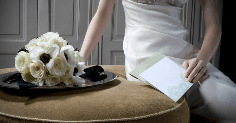 A woman in a wedding dress sitting on table, holding an envelope with a bouquet of flowers.